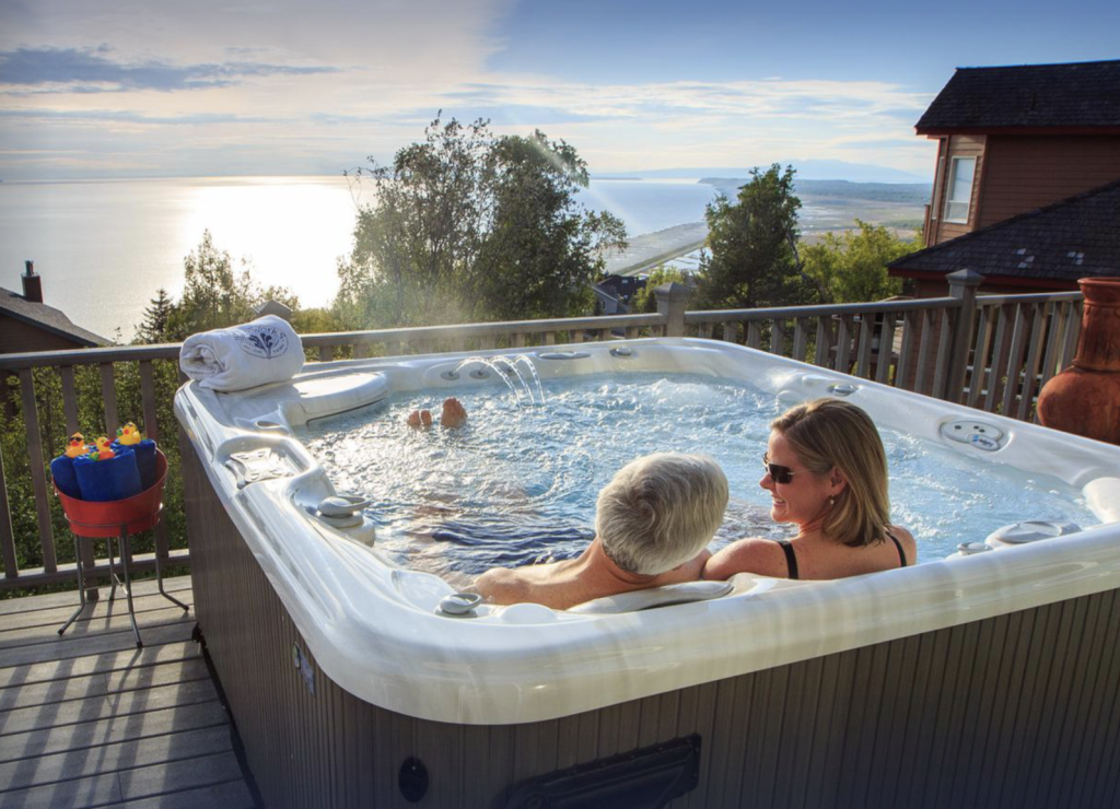 Two people relax in a hot tub on a deck overlooking a serene body of water with lush trees and clear skies.